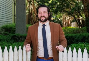 Executive Director of Visit St Francisville, Devan Corbello, in a suit outdoors by manicured bushes and a white fence.